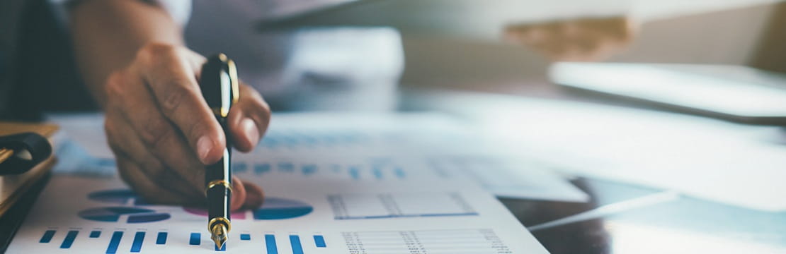 Hand pointing at printed charts with blue bar graphs while holding a tablet on a desk.